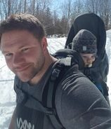 En mann smiler lett til kamera. På ryggen bærer han en baby i bæremeis.